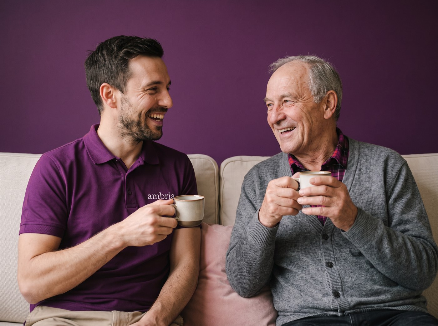 Ein Alltagshelfer und ein Klient sitzen mit Kaffeetassen auf dem Sofa und lachen miteinander