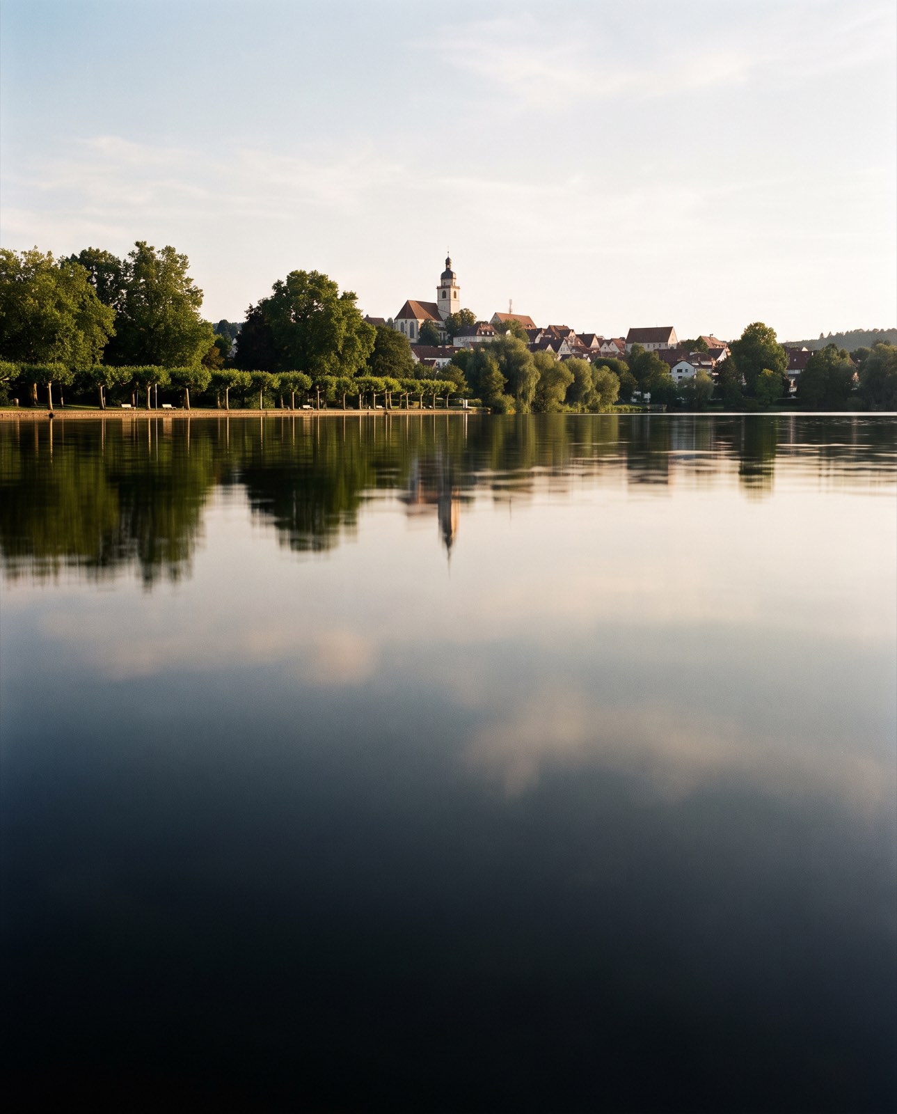 Ruhiger See mit Spiegelung im Wasser, am anderen Ufer Häuser und ein Kirchturm